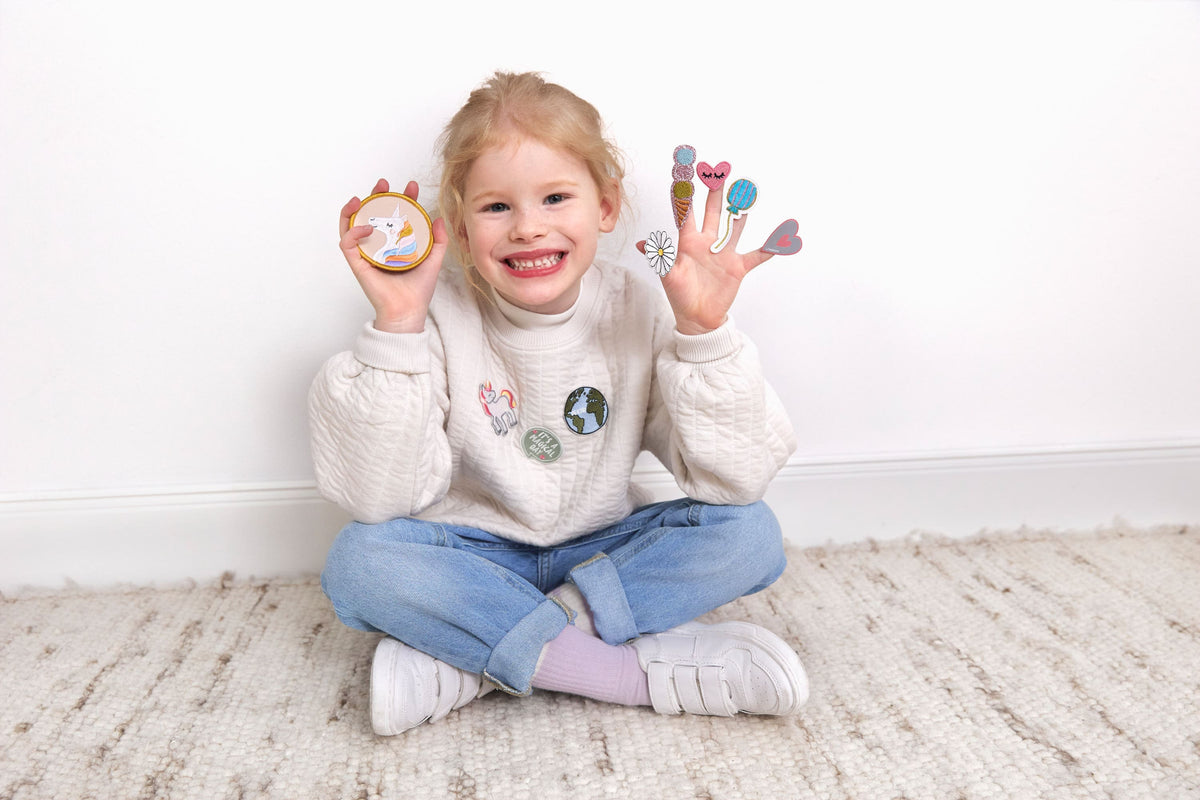 Lachendes Kind sitzt auf dem Boden und zeigt Laessig Patches & Anhaenger aus dem Einhorn Set, mit einem Einhorn-Patch in der linken Hand und verschiedenen Dekorationen in der rechten Hand.