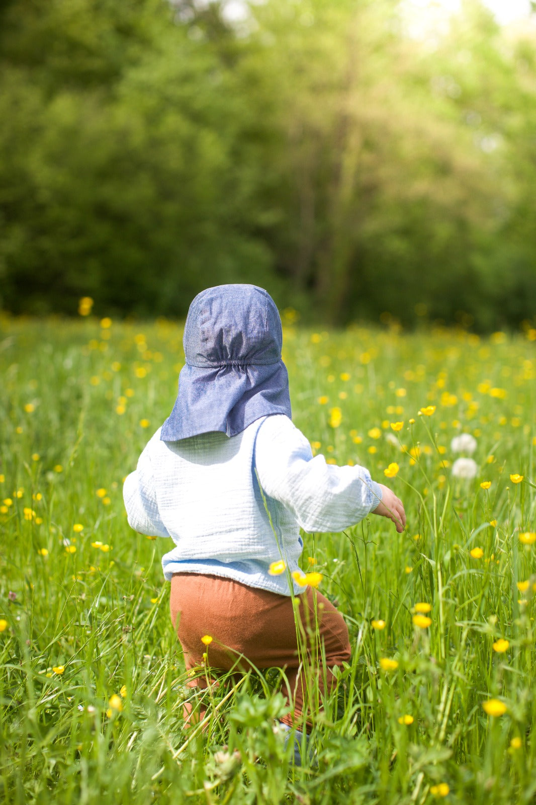 Kleinkind mit MP-Denmark Sonnenhut UV-50+ in Blau, spielend auf einer Sommerwiese mit gelben Blumen. Der Hut schützt die empfindliche Haut vor Sonnenstrahlung und bietet dank Nackenschutz optimalen Schutz.